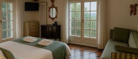A bedroom with a large bed, a green armchair, a wooden floor, and a TV on the wall.