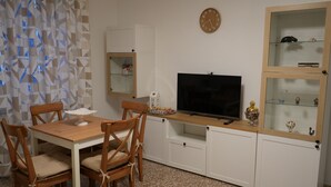 A compact living space with a wooden dining table and chairs, a flat-screen TV, a small cabinet with glass doors, and a wall clock.