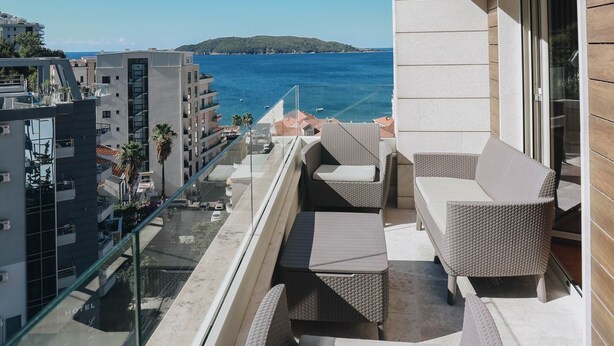 Een balkon met rieten meubels, een glazen hekwerk en uitzicht op zee en gebouwen.