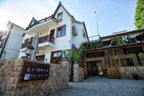 A two-story building with a stone entrance, a balcony on the upper floor, and a covered area with plants.