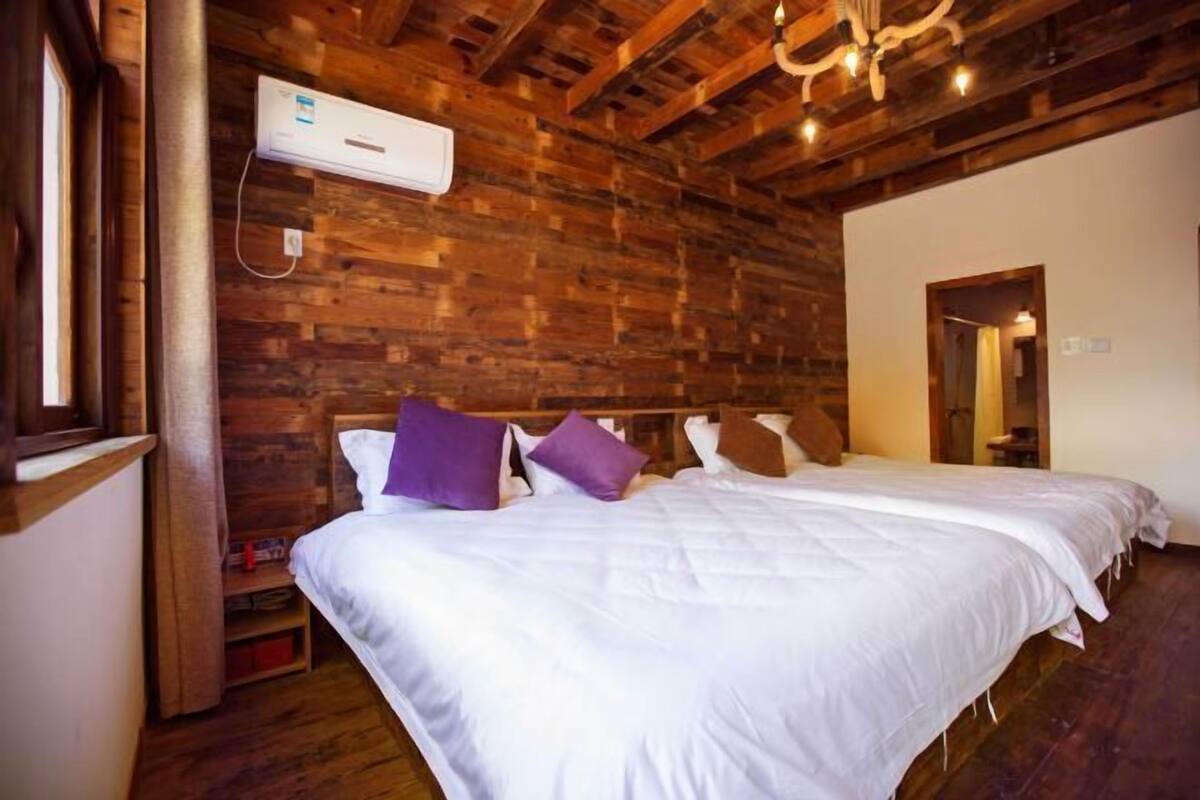 Una habitación con techo de madera, cama con ropa de cama blanca y almohadas moradas, y un aire acondicionado instalado en la pared.
