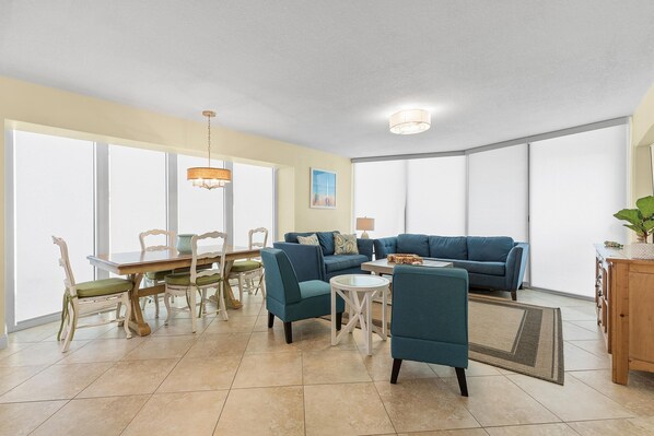 A spacious living room with a large window, a blue sofa, a dining table with chairs, and a small round table. - Golden Arms 101 (New Smyrna Beach)