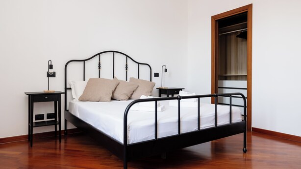 A single bed with a metal frame, a wooden floor, a nightstand with a lamp, and a wooden wardrobe.