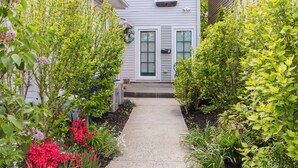 A pathway leading to a house with a front door and windows, surrounded by lush greenery and flowering plants.