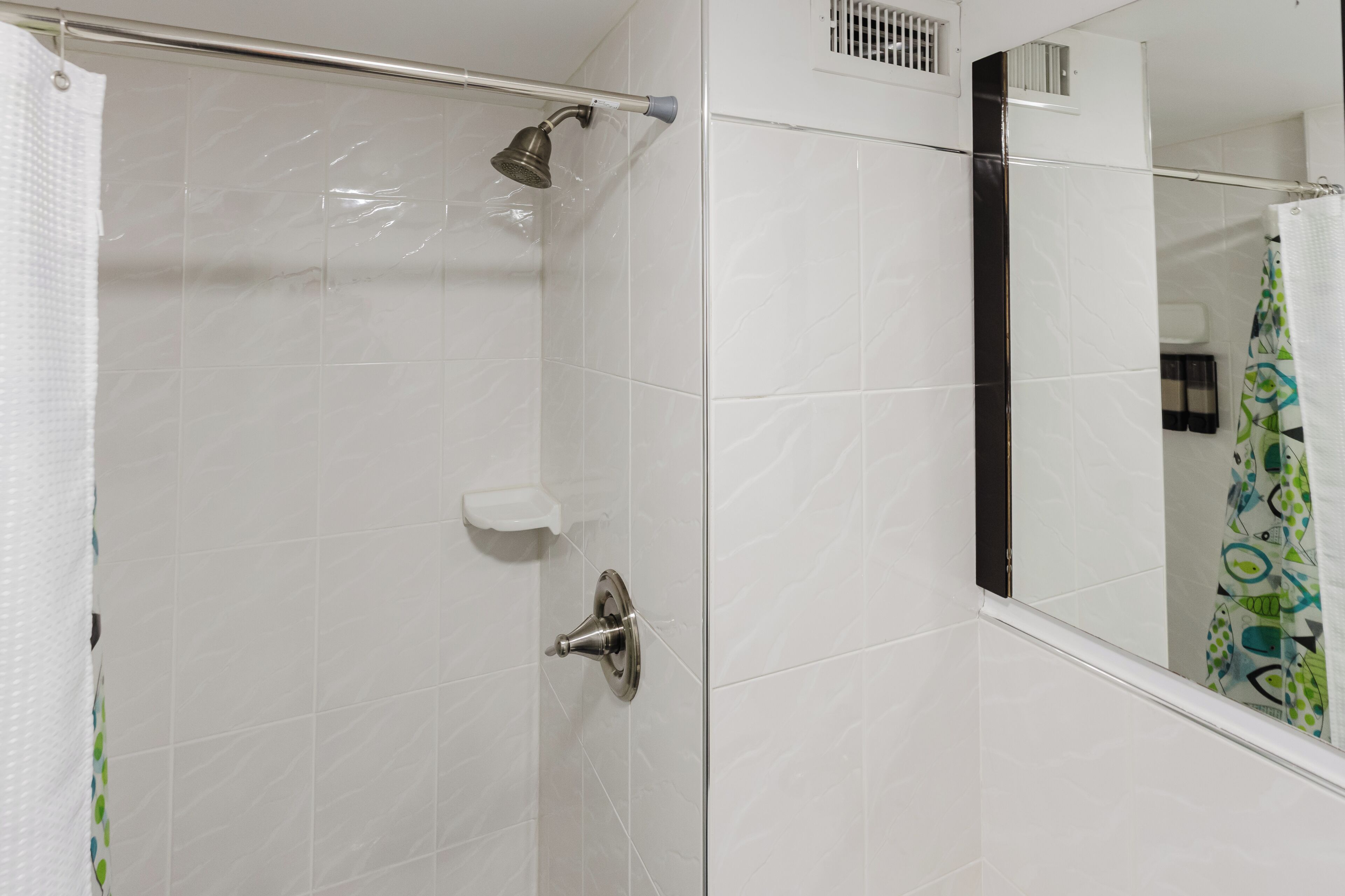 Ein Badezimmer mit weißen Fliesen, einem Duschvorhang, einer Handbrause, einer kleinen Ablage und einem Spiegel, der einen farbenfrohen Duschvorhang reflektiert.