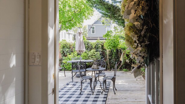 An open door leading to a patio with a checkered mat, metal chairs, a table, and a hanging plant.