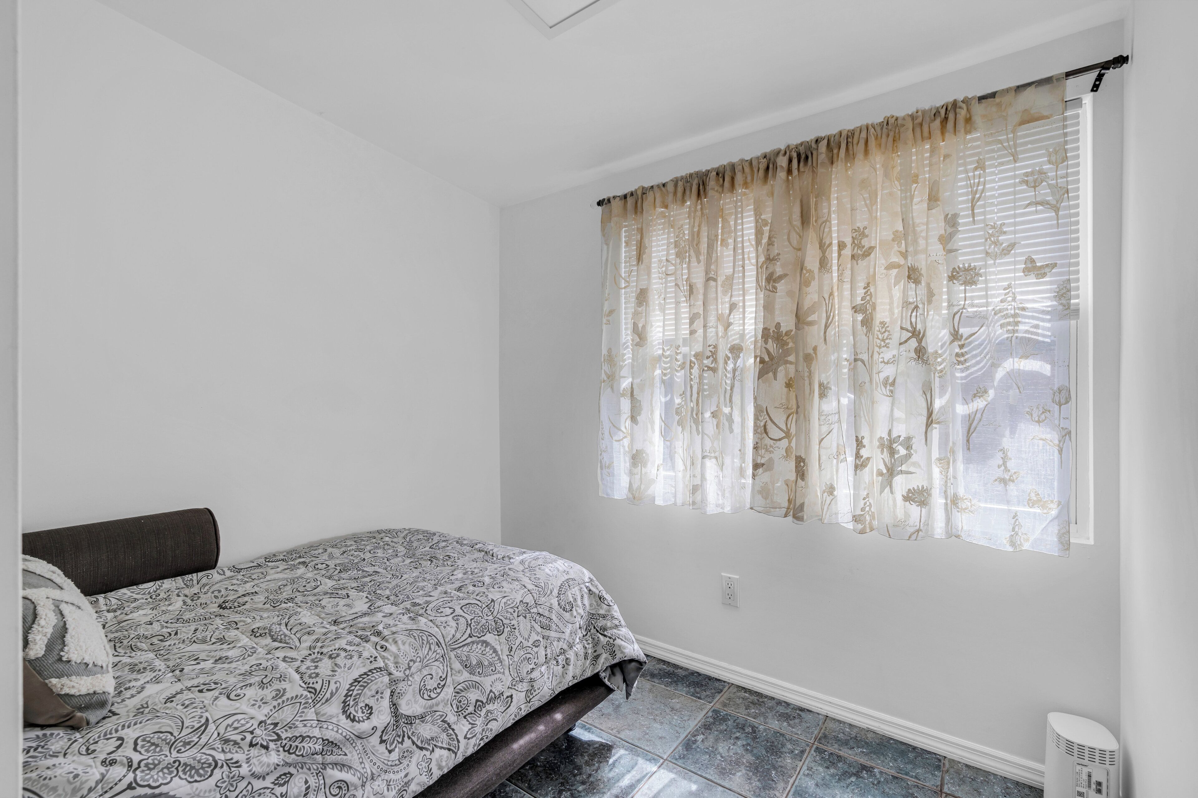 A bedroom with a bed, a window with floral curtains, and a white wall.