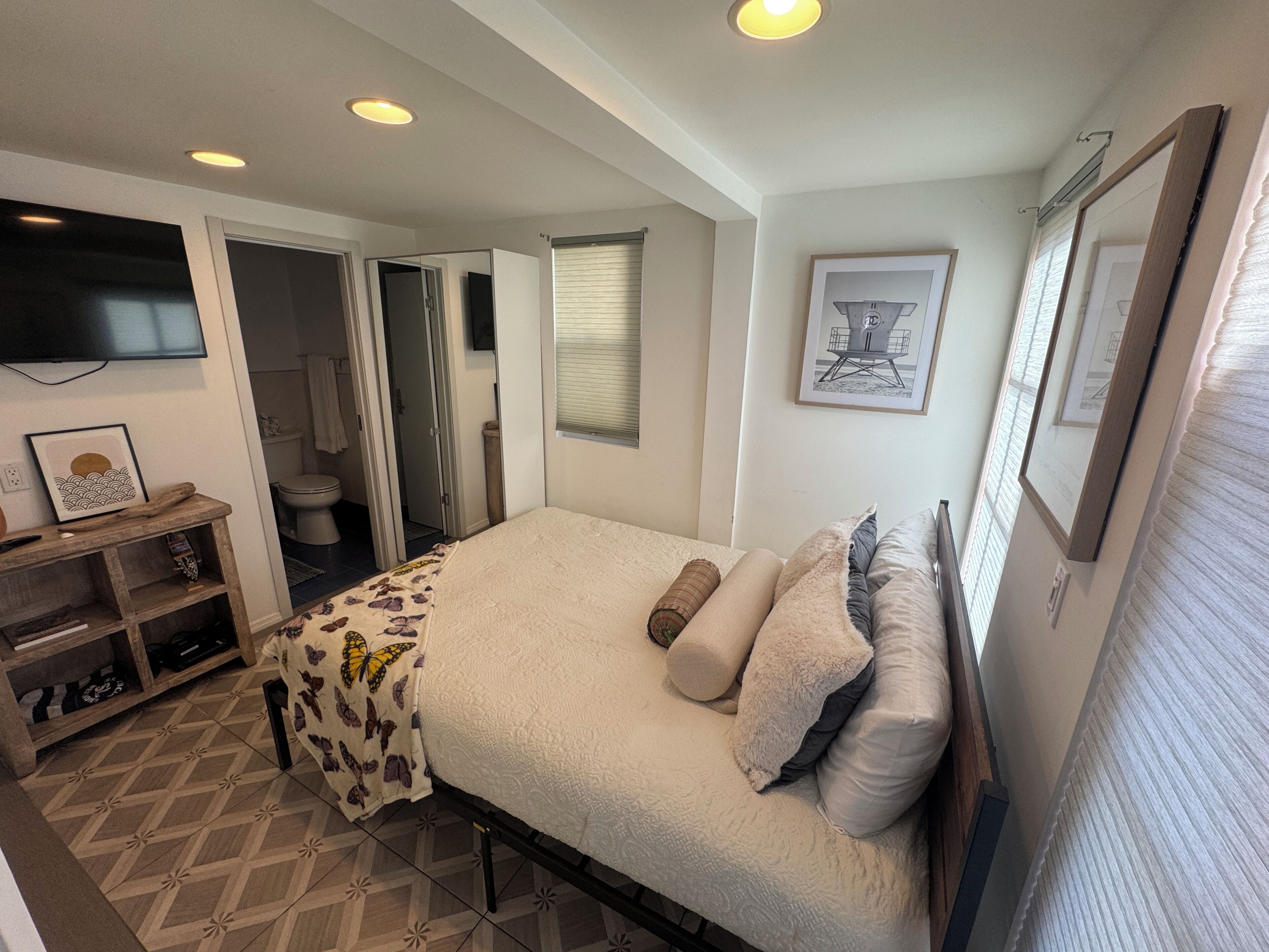 Un dormitorio con cama, televisión, un cuadro enmarcado y baño visible a través de una puerta abierta.