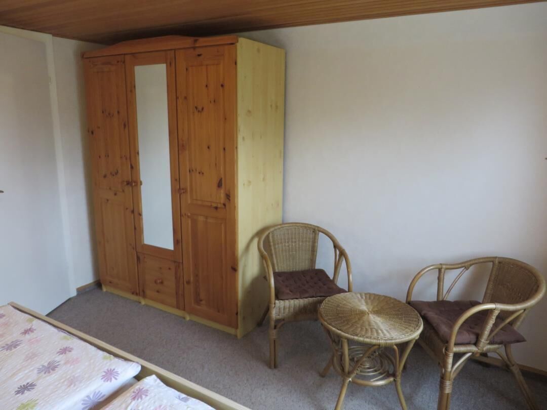 A bedroom with a wooden wardrobe, two wicker chairs, and a bed with a floral bedspread.