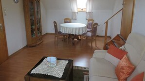 A living room with a round table, chairs, a sofa, a cabinet, and a window with curtains. - NEW! Vacation apartment directly on the dike (Dornum)
