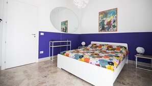 A bedroom with a bed, a white door, a mirror, a round mirror, a colorful painting, and a white pendant light. - Camera con bagno in centro città (Reggio Calabria)