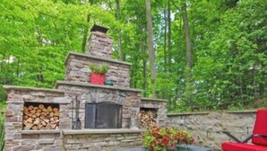 An outdoor stone fireplace with logs, surrounded by red chairs and a flower arrangement.