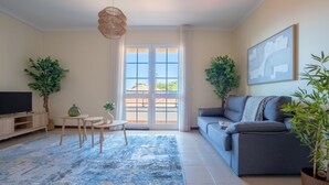 Ein Wohnzimmer mit Sofa, Fernseher, kleinem Tisch, Sesseln und Topfpflanzen.