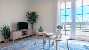 Ein Wohnzimmer mit Fernseher auf Holzständer, Topfpflanze, Vase und Blick durch ein großes Fenster.