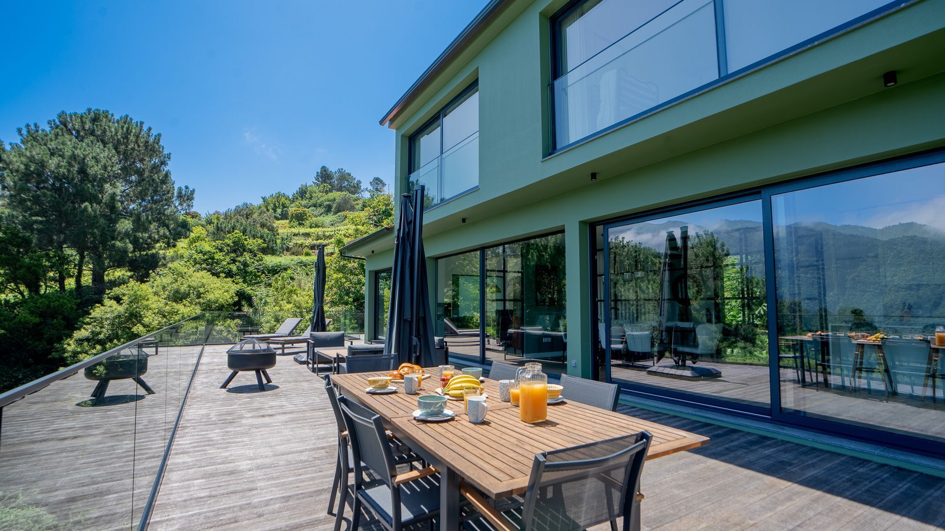 A modern outdoor dining area with a wooden table set for breakfast, overlooking a lush green landscape and a mountain range.