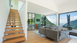 Ein modernes Interieur mit einer Holztreppe, einem grauen Sofa und einem Essbereich mit Blick auf eine gebirgige Landschaft.