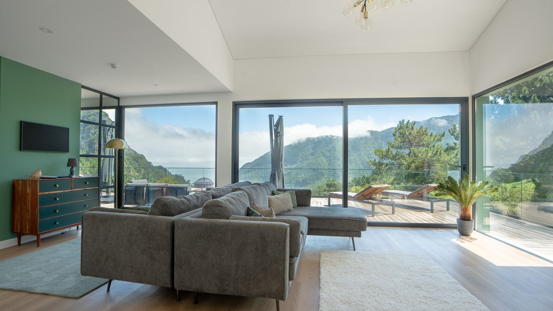 A modern living room with a large sofa, a wooden cabinet, and a TV on the wall. There are large windows offering a view of mountains and greenery.