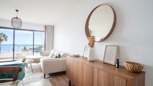 A modern living room with a large window overlooking the sea, a round mirror, a wooden cabinet, and a teal armchair. - Hygge Madeira by LovelyStay (Canico)