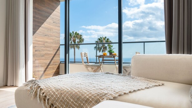 A modern living room with a large window, a sofa, a coffee table, and a view of palm trees and the ocean. - Hygge Madeira by LovelyStay (Canico)