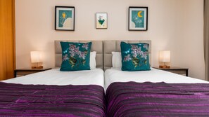 A hotel room with two beds, a nightstand with a lamp, and three framed artworks on the wall.