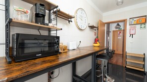 A kitchenette with a black microwave, a coffee machine, and a toaster. There is a wooden shelf with jars and a clock on the wall.
