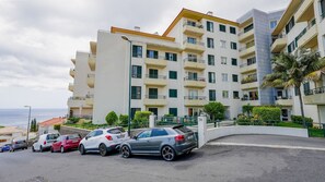 Un immeuble résidentiel à plusieurs étages, doté de balcons, d’un espace de stationnement où sont garées plusieurs voitures, et offrant une vue sur la mer.