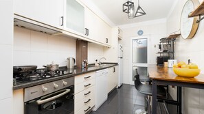 A modern kitchen with a gas stove, oven, and a coffee maker. There is a dining area with a wooden table and a yellow bowl of fruit. - Delmar by LovelyStay (Caniço)