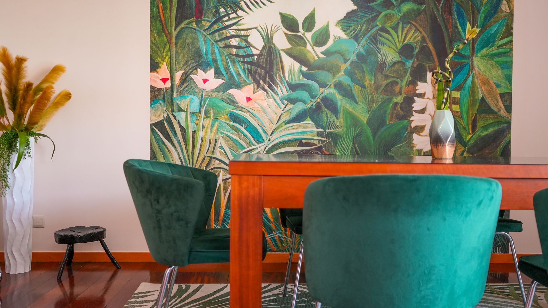 Un coin repas avec une table en bois et des chaises de couleur turquoise, un vase rempli de fleurs et un tableau aux motifs tropicaux accroché au mur.
