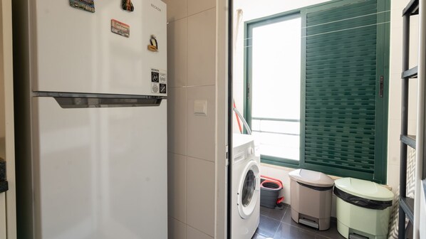 A compact kitchen with a refrigerator, a washing machine, and a trash bin.