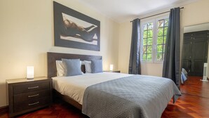 Une chambre à coucher comprenant un lit, des tables de chevet, une commode, une fenêtre avec des rideaux et un tableau au mur.