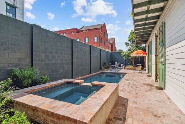 Ein Pool mit Steinrand, eine Terrasse mit Gartenmöbeln und ein rotes Gebäude im Hintergrund.
