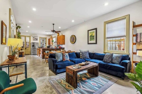 A spacious living room with a blue sofa set, a wooden coffee table, and a patterned rug. A dining area with a table and chairs is visible in the background.