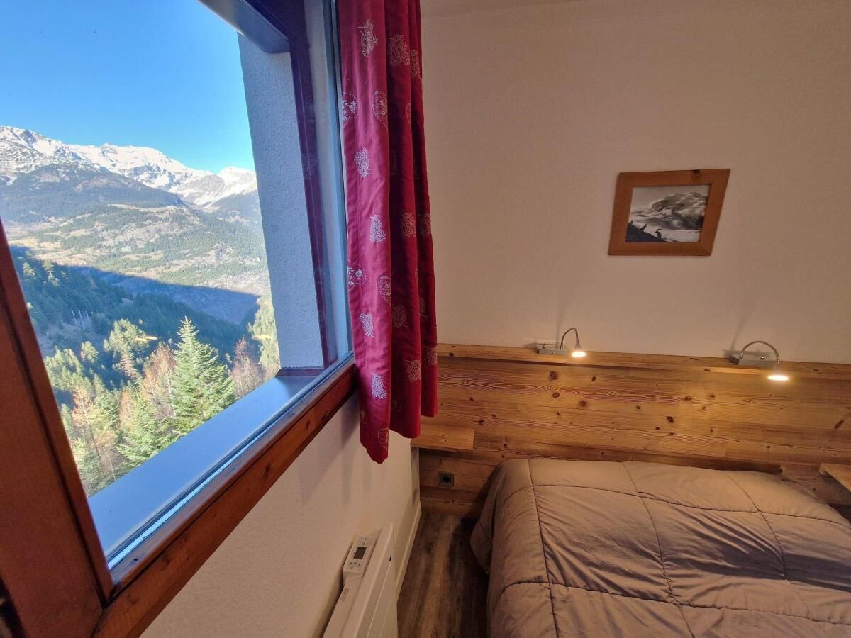 Een slaapkamer met een houten hoofdbord, een bed met een beige deken, een raam met uitzicht op bergen en bomen, en een klein fotolijstje aan de muur.