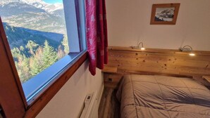 A bedroom with a wooden headboard, a bed with a beige blanket, a window with a view of mountains and trees, and a small framed picture on the wall.
