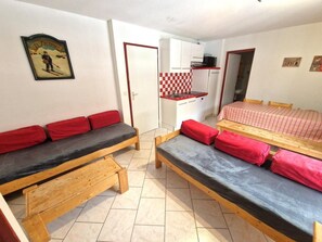 Habitación compacta con dos camas de madera, ropa de cama roja y negra, una kitchenette pequeña y un salpicadero a cuadros.