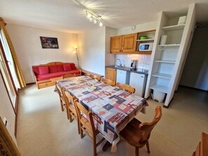 Espaço compacto com mesa de jantar, cadeiras de madeira, sofá vermelho, cozinha com armários e micro-ondas, e uma pequena estante.