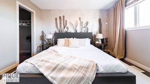 A bedroom with a bed, a nightstand, a lamp, a cactus wall decoration, and a window with curtains. - Chic 6BDRM Getaway - KingBed - Patio - BBQ - Garage - Wifi (Edmonton)