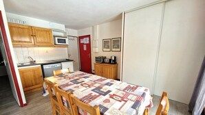 Uma cozinha compacta com armários de madeira, micro-ondas e um frigorífico pequeno. Uma mesa de jantar com toalha quadriculada está posta para quatro pessoas.