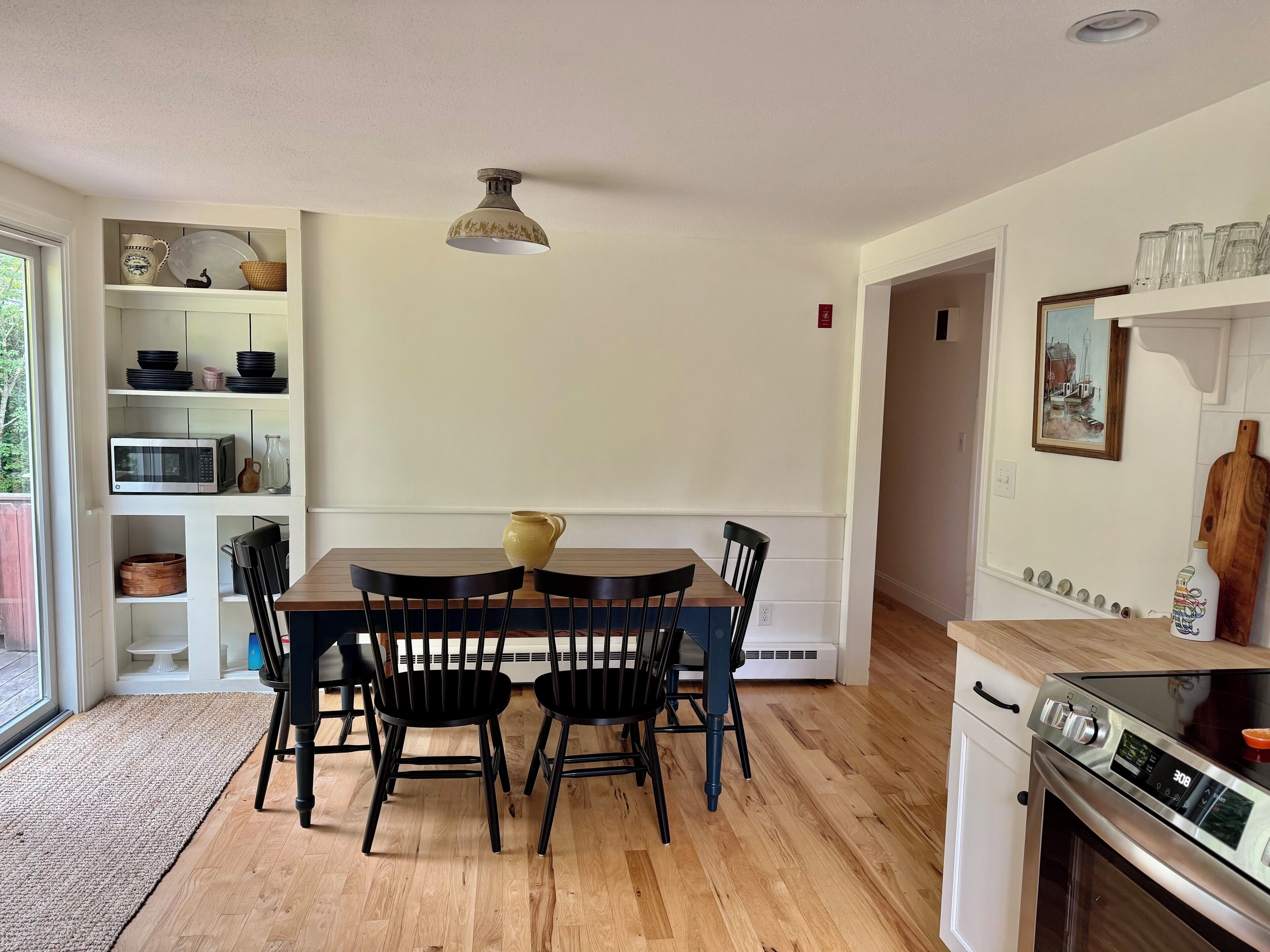 A dining area with a wooden table and chairs, a hanging light fixture, and a kitchen with a stove and microwave.