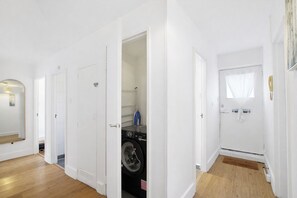 A clean, modern apartment with white walls and wooden flooring. It includes a laundry room with a washing machine and an ironing board.