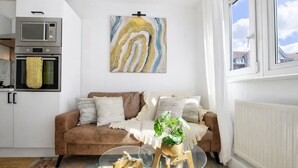 A living room with a brown sofa, a glass coffee table, a painting, and a kitchen area with a microwave. - Central Square Apartments (London)