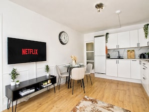 A modern living room with a flat-screen TV, a dining area with chairs, a small table, and a kitchen with white cabinets.