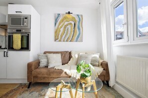 A living room with a brown sofa, a glass coffee table with a plant, a kitchen with a microwave, and a painting on the wall.