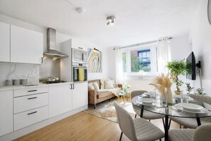 A modern kitchen with white cabinetry, a stove, and a microwave. There is a dining area with a glass table and chairs.