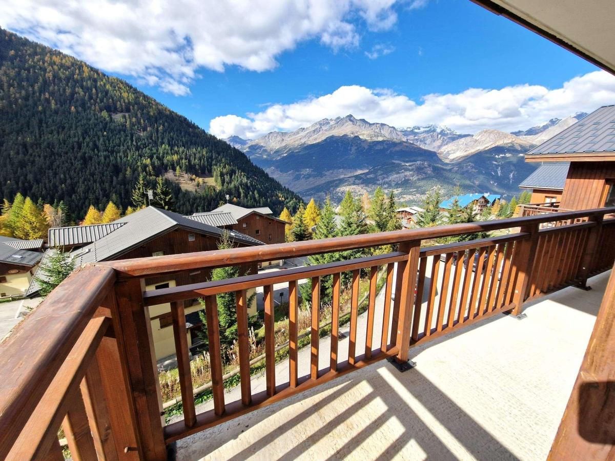 A wooden balcony with a view of a mountainous landscape, including houses and trees.