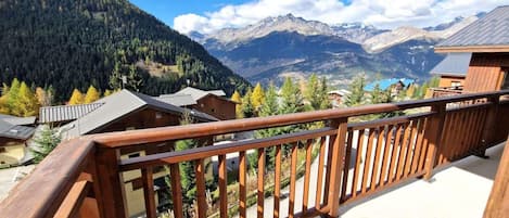 A wooden balcony with a view of a mountainous landscape, including houses and trees.