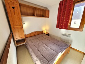 Una habitación compacta con cama, armario de madera, mesita de noche con lámpara y ventana con cortinas rojas.