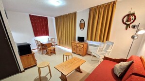 Un salon avec un canapé rouge, une table basse en bois et des fauteuils. On y voit un téléviseur posé sur un meuble en bois, ainsi qu’une petite table avec une chaise. Un rideau rouge et un élément de décoration murale sont visibles.