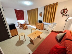 A living room with a red sofa, wooden coffee table, and chairs. There is a TV on a wooden cabinet, and a small table with a chair. A red curtain and a wall decoration are visible. - 100m from ski slopes, 2nd floor, balcony, ski locker, 36m², Valfréjus (Valfréjus)