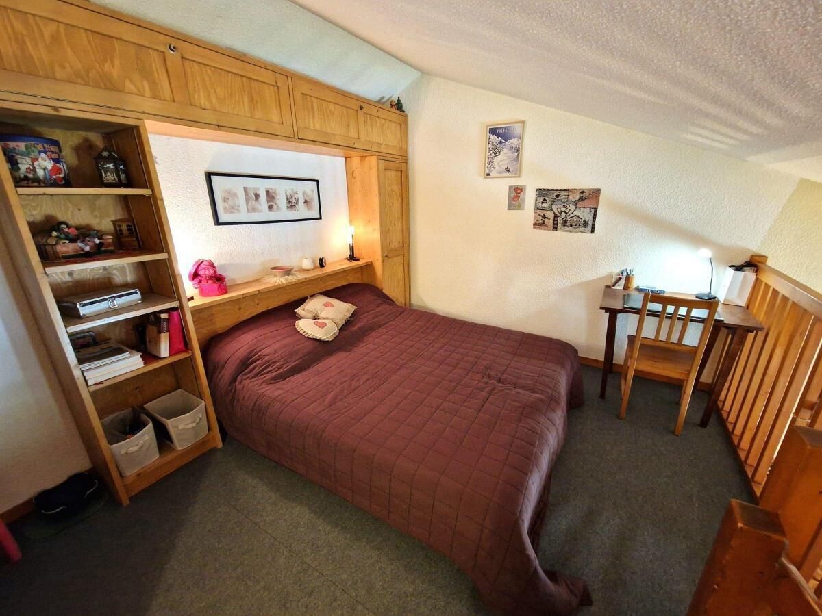 A small, cozy bedroom with a single bed, a wooden wardrobe, a desk with a chair, and a small table with a lamp.